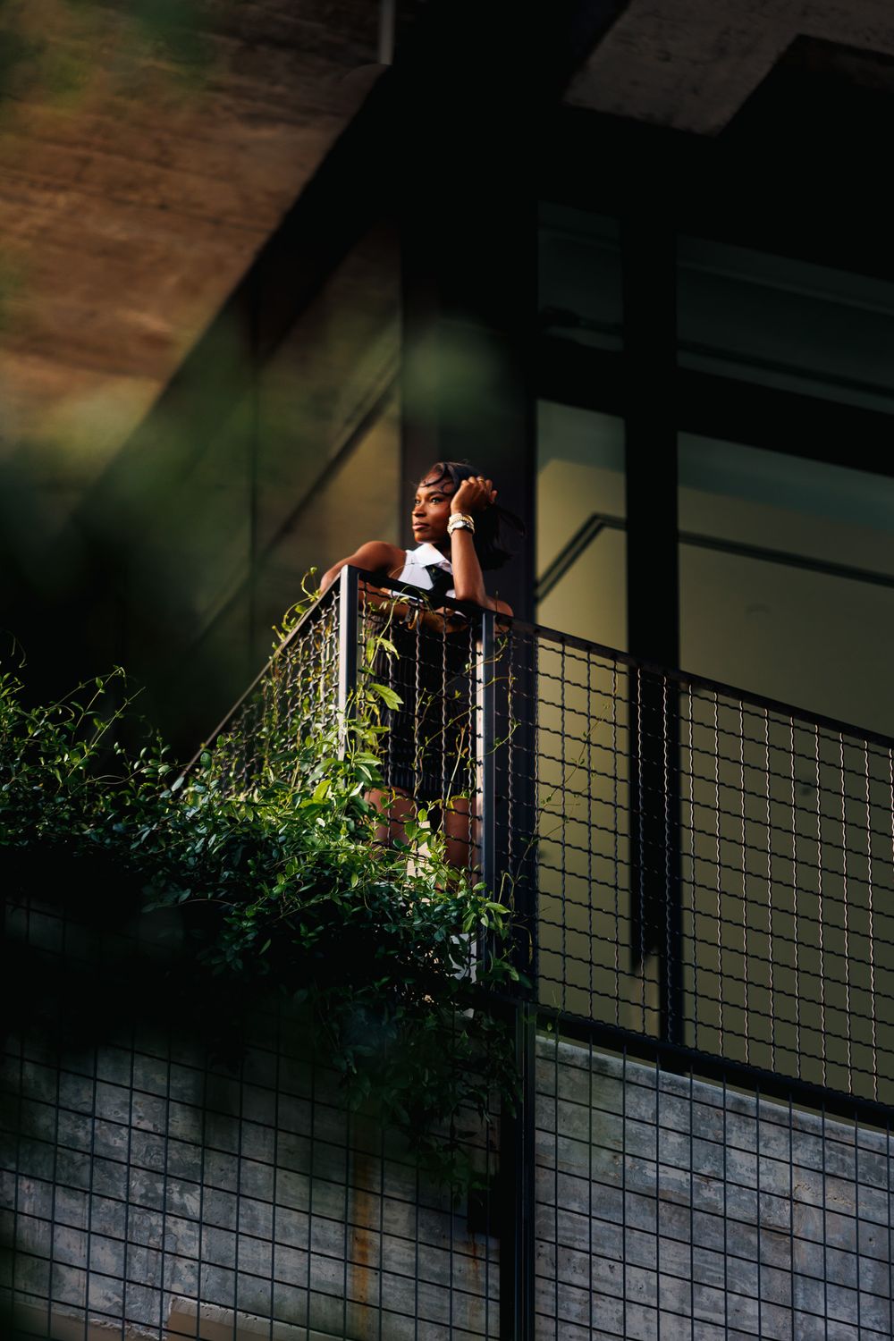 A person stands on a dark balcony with metal railings at night, surrounded by greenery and dramatic lighting.