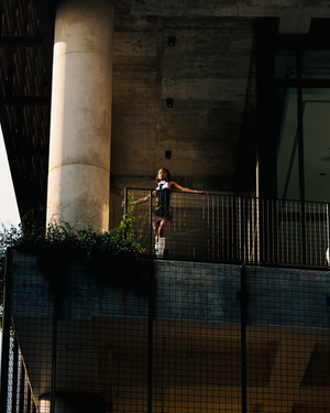 A silhouetted figure stands on a bridge against a dramatic backlit urban setting with concrete columns.