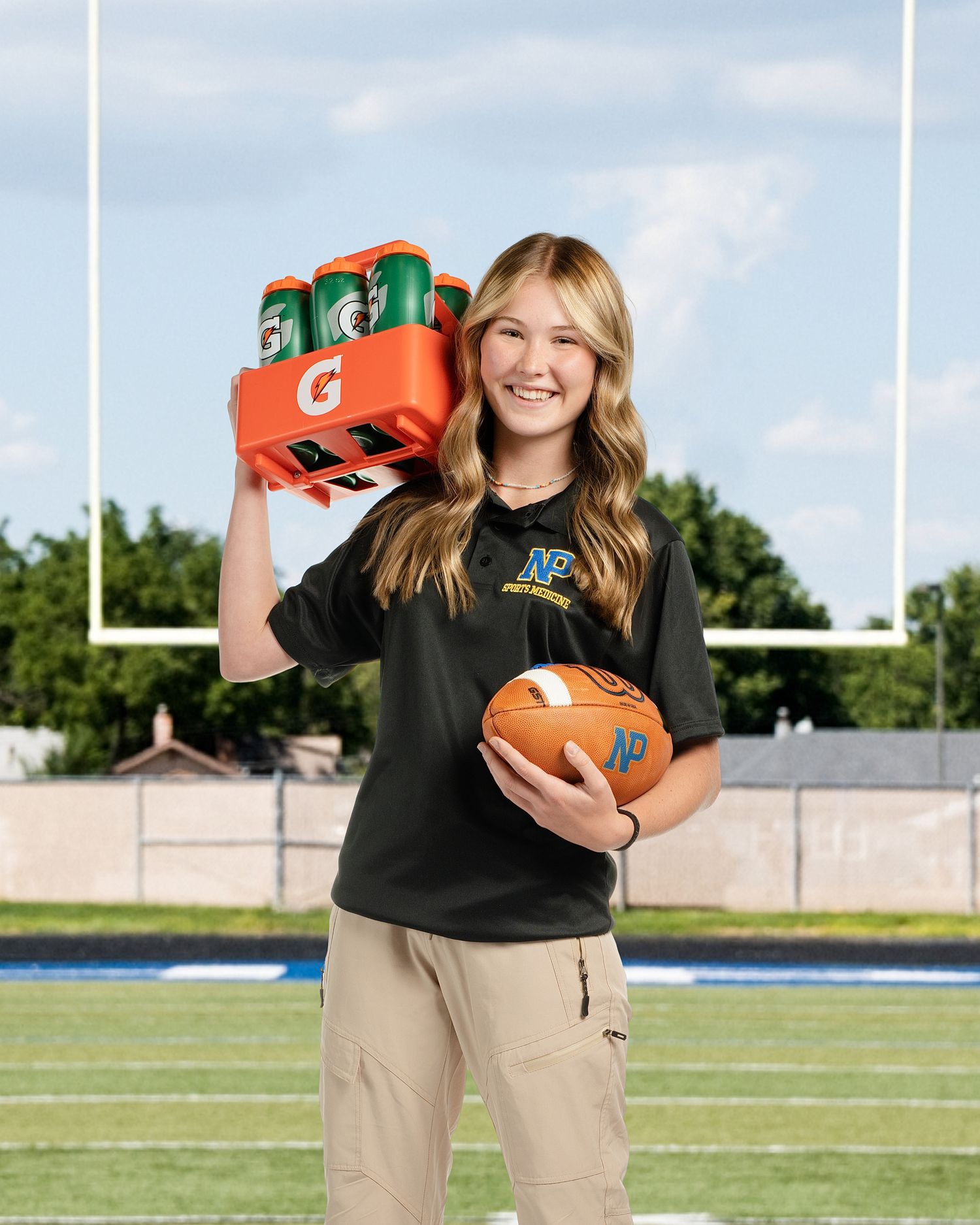 Athletic trainer poses on football field with sports equipment and Gatorade bottles.