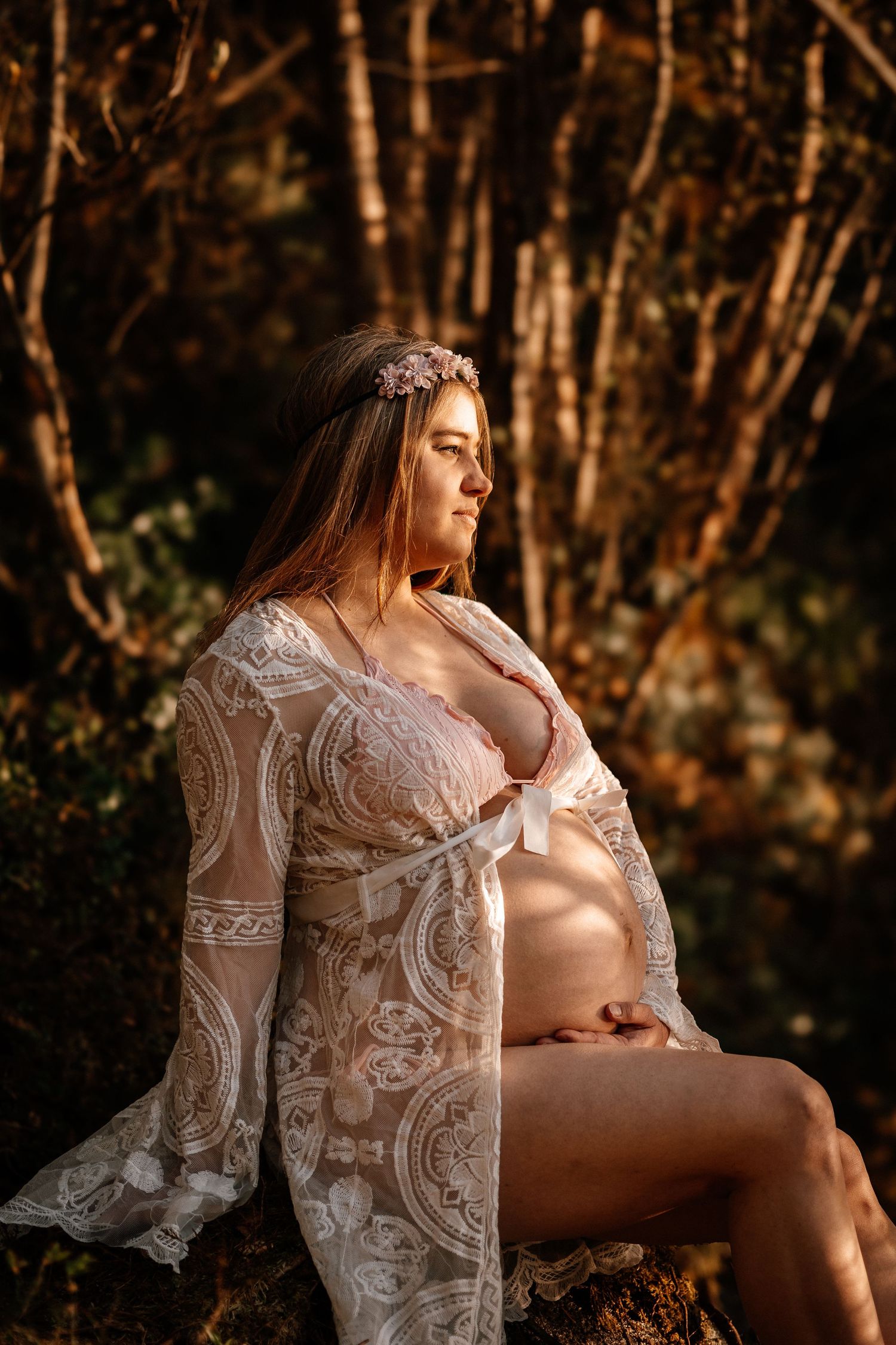 Art photography of a maternity portrait with delicate lace robe in warm sunset lighting among trees and woodland.