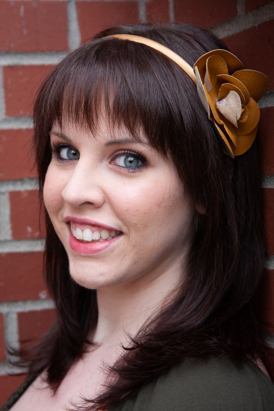 A close-up portrait against a brick wall showing dark hair with bangs, a gold flower headband, and warm smile.