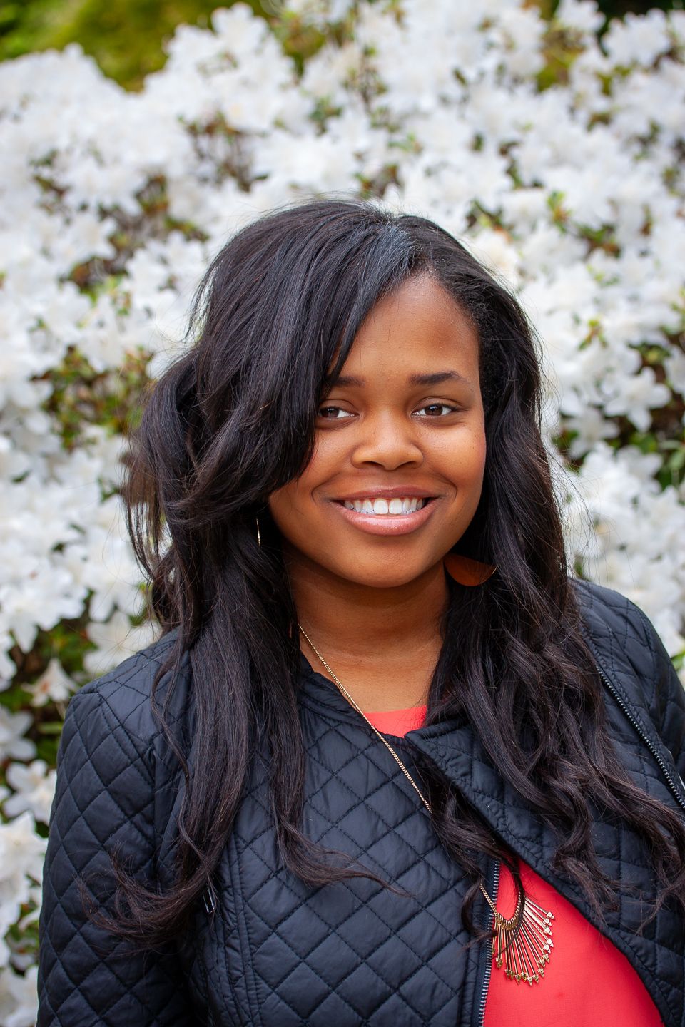 Portrait of someone wearing a black quilted jacket and red top smiling outdoors with white flowering trees in background.