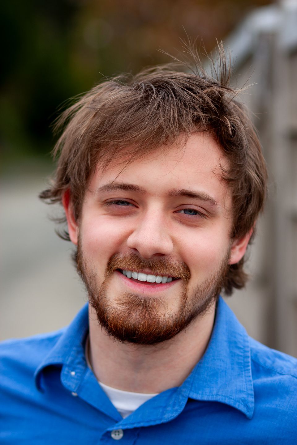 Portrait of smiling young person with brown hair and beard wearing a blue collared shirt against blurred background.