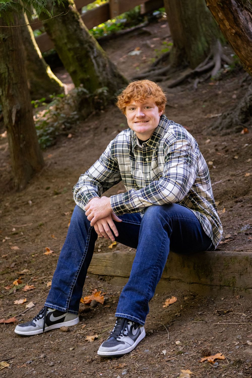 A person in a plaid flannel shirt and jeans sits casually on a wooden log in a wooded outdoor setting.