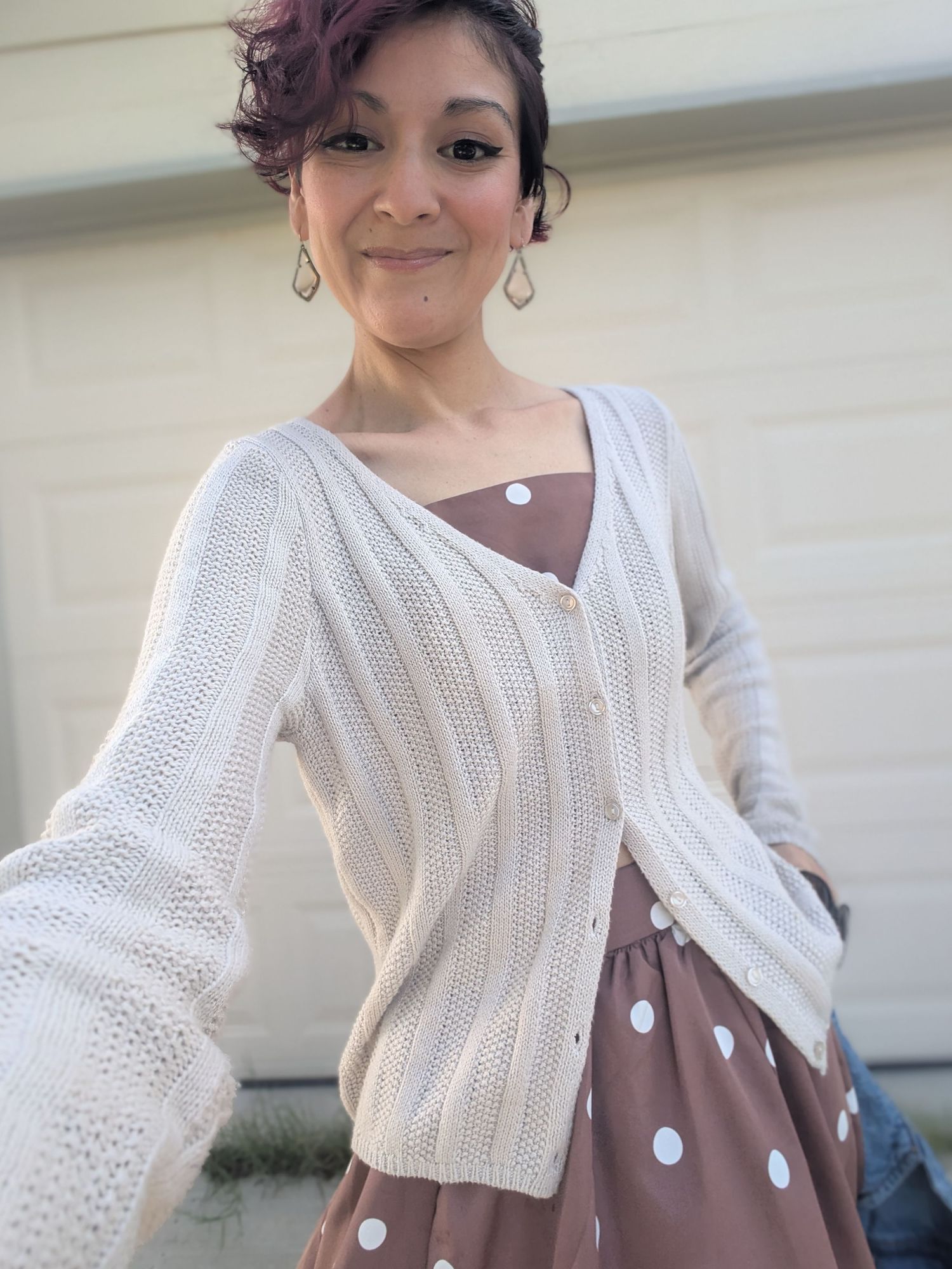 Woman in white knit cardigan and brown sleeveless dress standing against light colored wall.