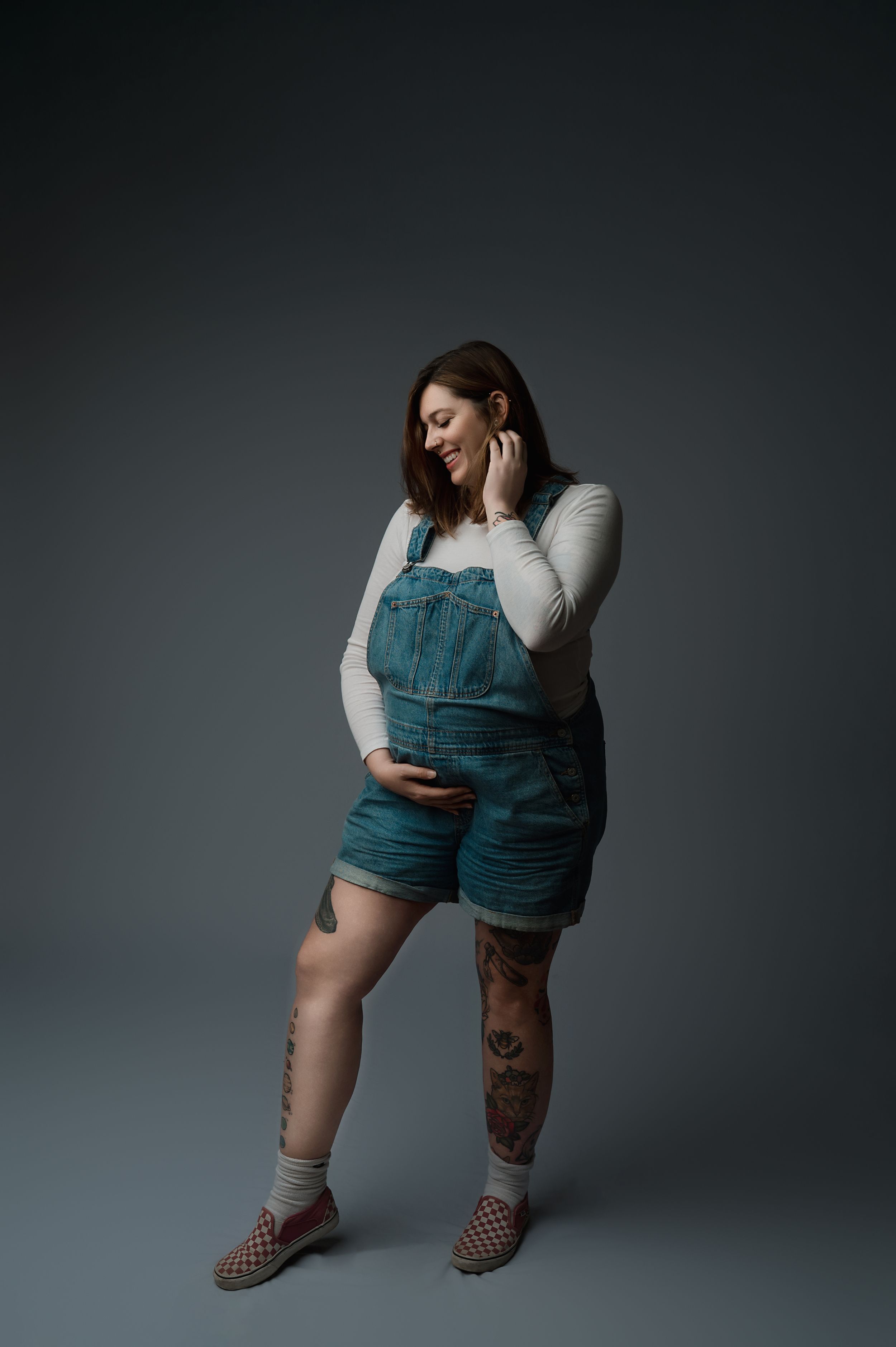Expecting figure stands in denim overalls and white top against gray backdrop, with tattoos visible on leg.