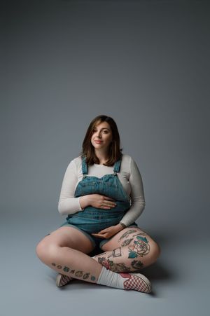 A person wearing teal overalls and white shirt sits cross-legged on the floor with visible leg tattoos against a grey backdrop.