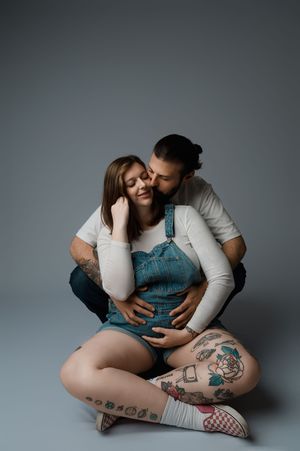 A tender moment between a couple on a gray background, featuring denim overalls and visible tattoos.
