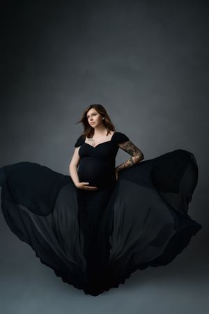 Dramatic maternity portrait of a woman in flowing black gown against dark background with billowing fabric movement.