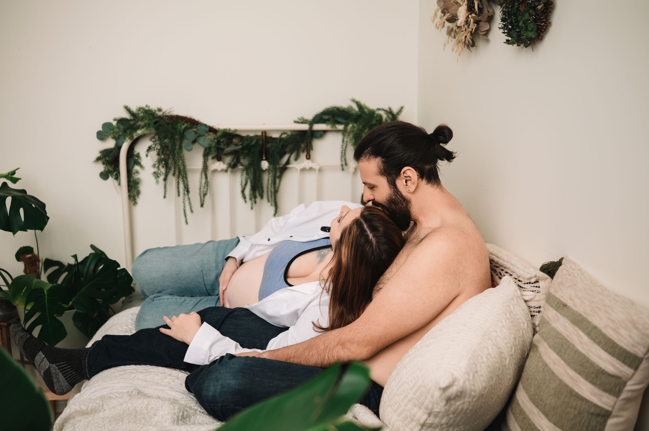 A couple cuddles together on a couch surrounded by plants and greenery in a cozy home setting.
