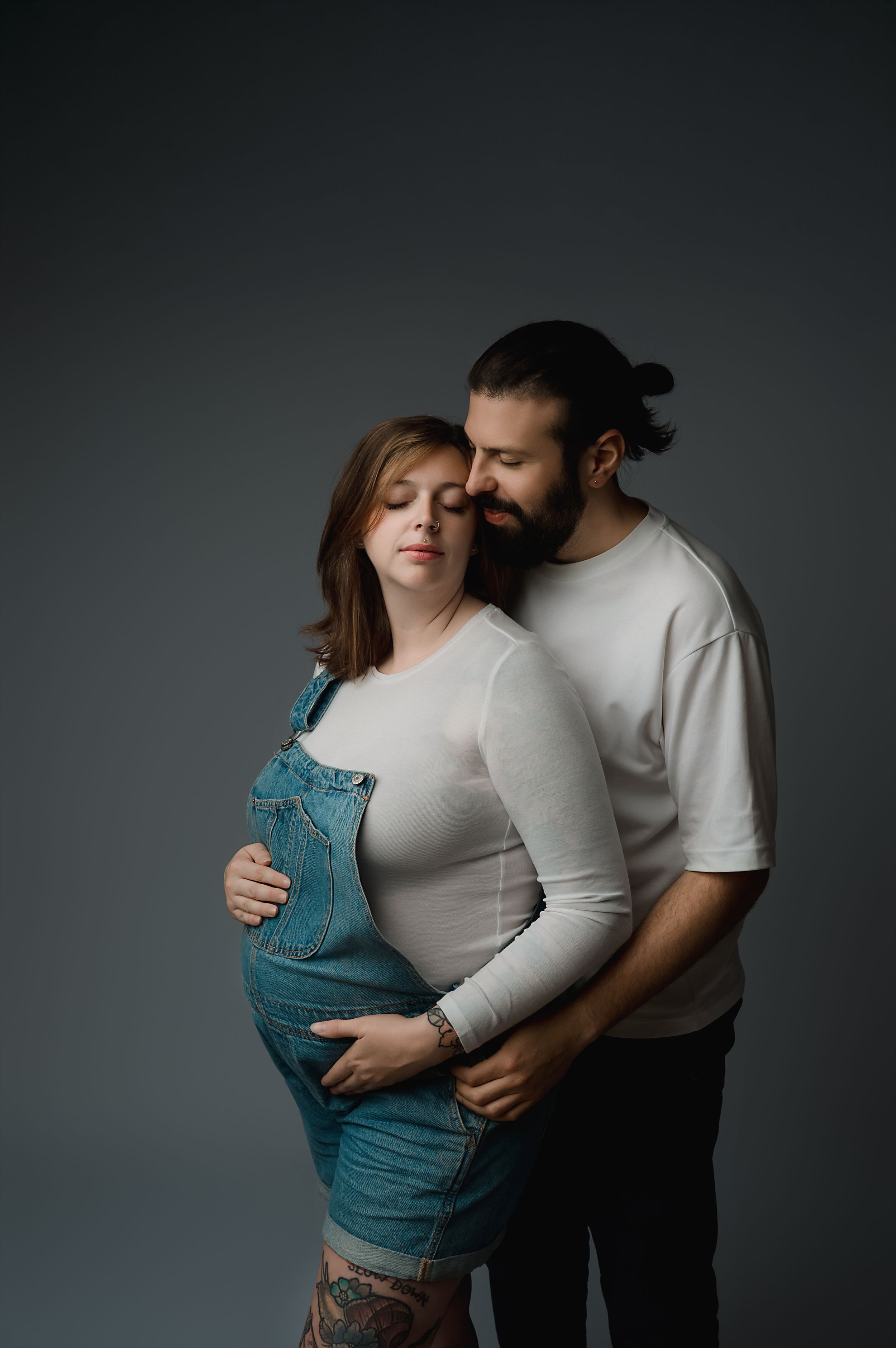 A tender maternity portrait with a couple embracing against a dark gray backdrop, wearing casual clothing.