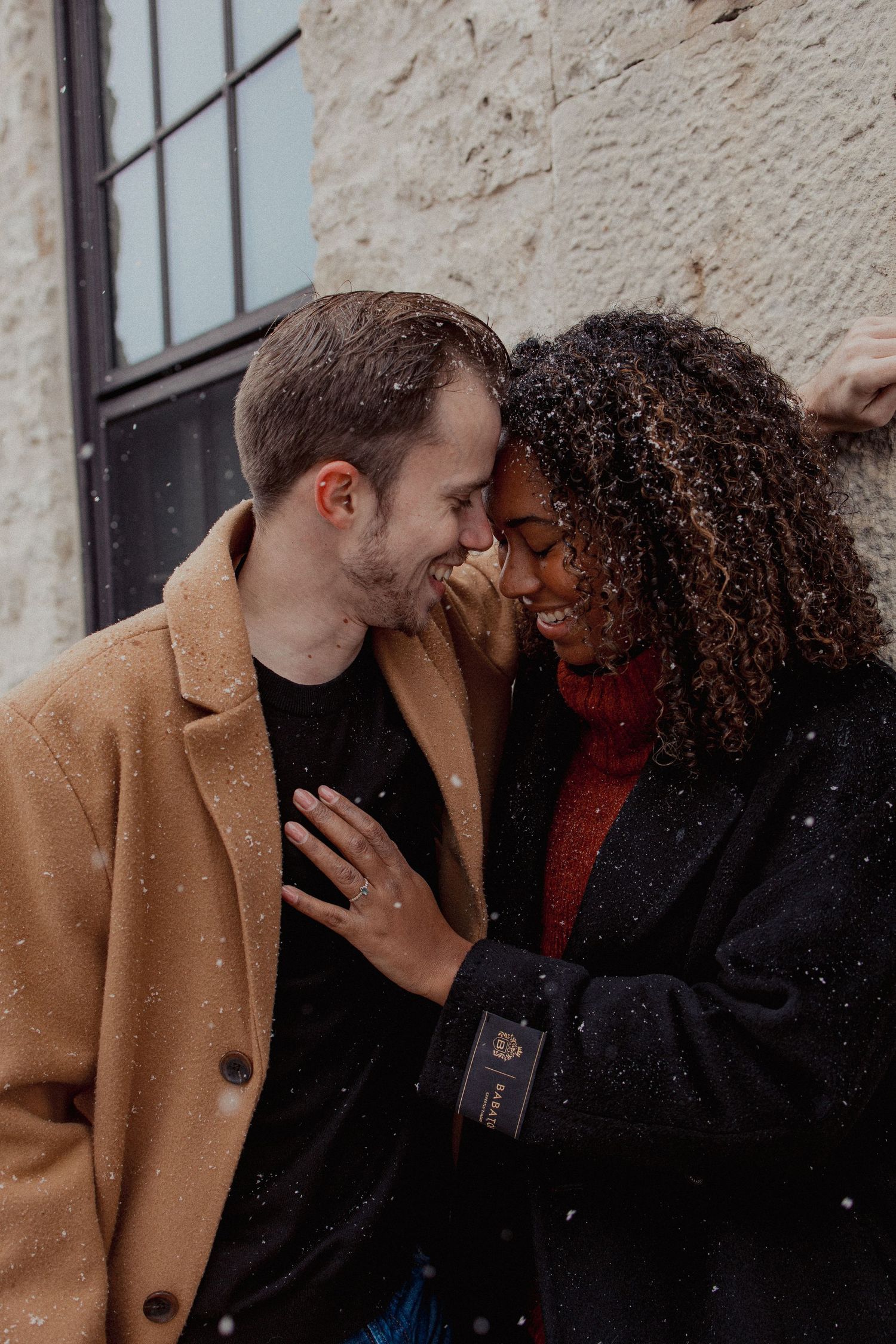 Interracial couple laughs and touches foreheads romantically in the snow against a stone wall.