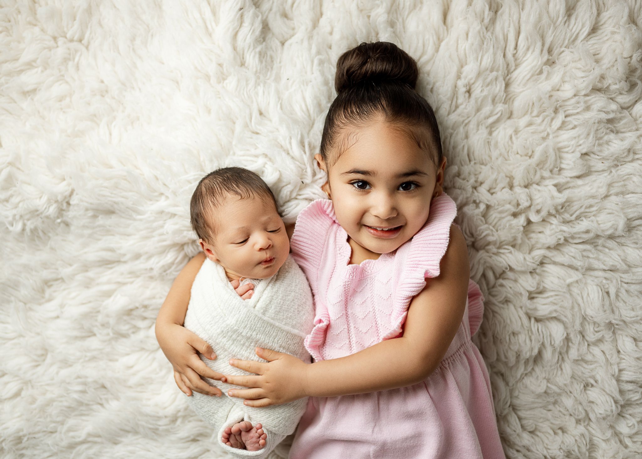 A young sibling in a pink dress holding a sleeping baby while lying on a white fur rug together.