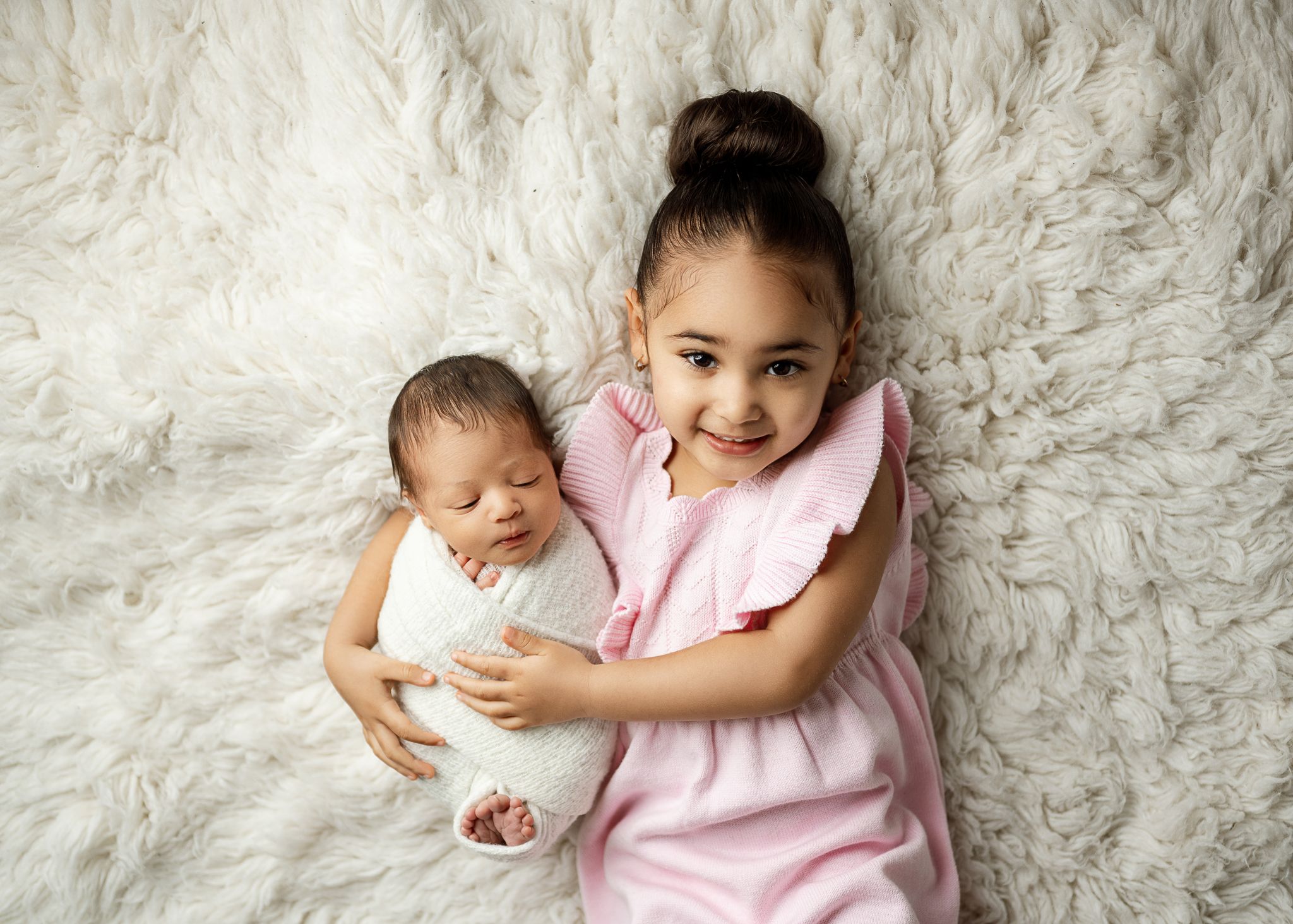 A little girl in a pink dress tenderly holds a sleeping newborn baby while lying on a soft white fur backdrop.