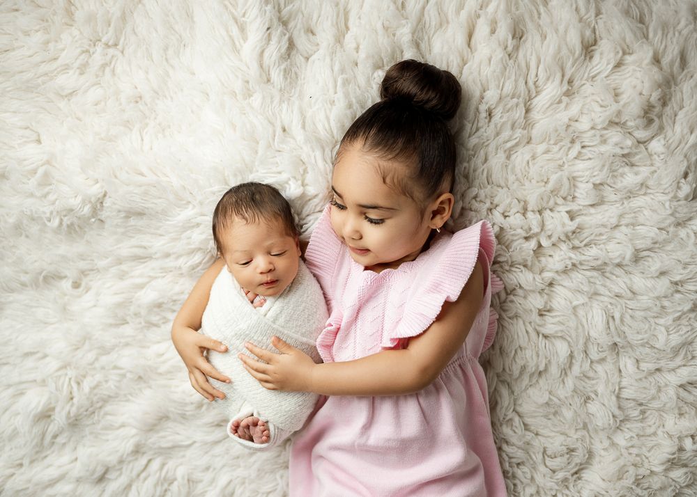 A young child in a pink dress holds a newborn baby while sitting on a soft white furry backdrop.