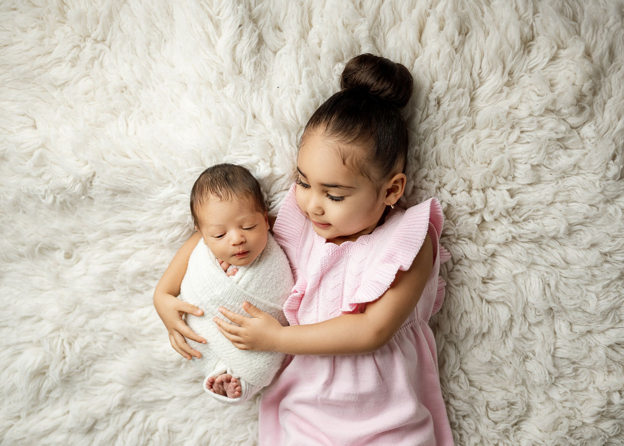 A young child in a pink dress holds a newborn baby while sitting on a soft white furry backdrop.