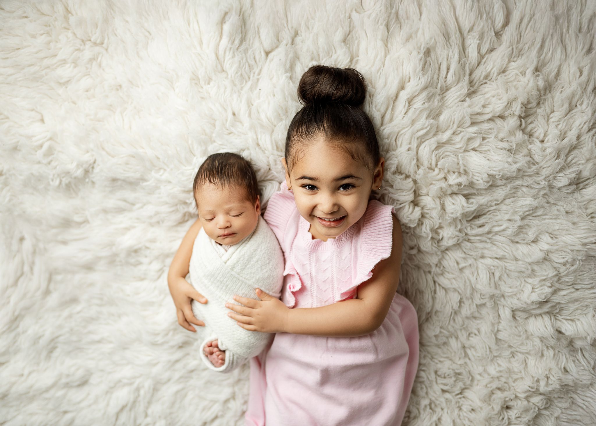 Two young children lying on white furry rug, one in white onesie and other in pink shirt smiling at camera.