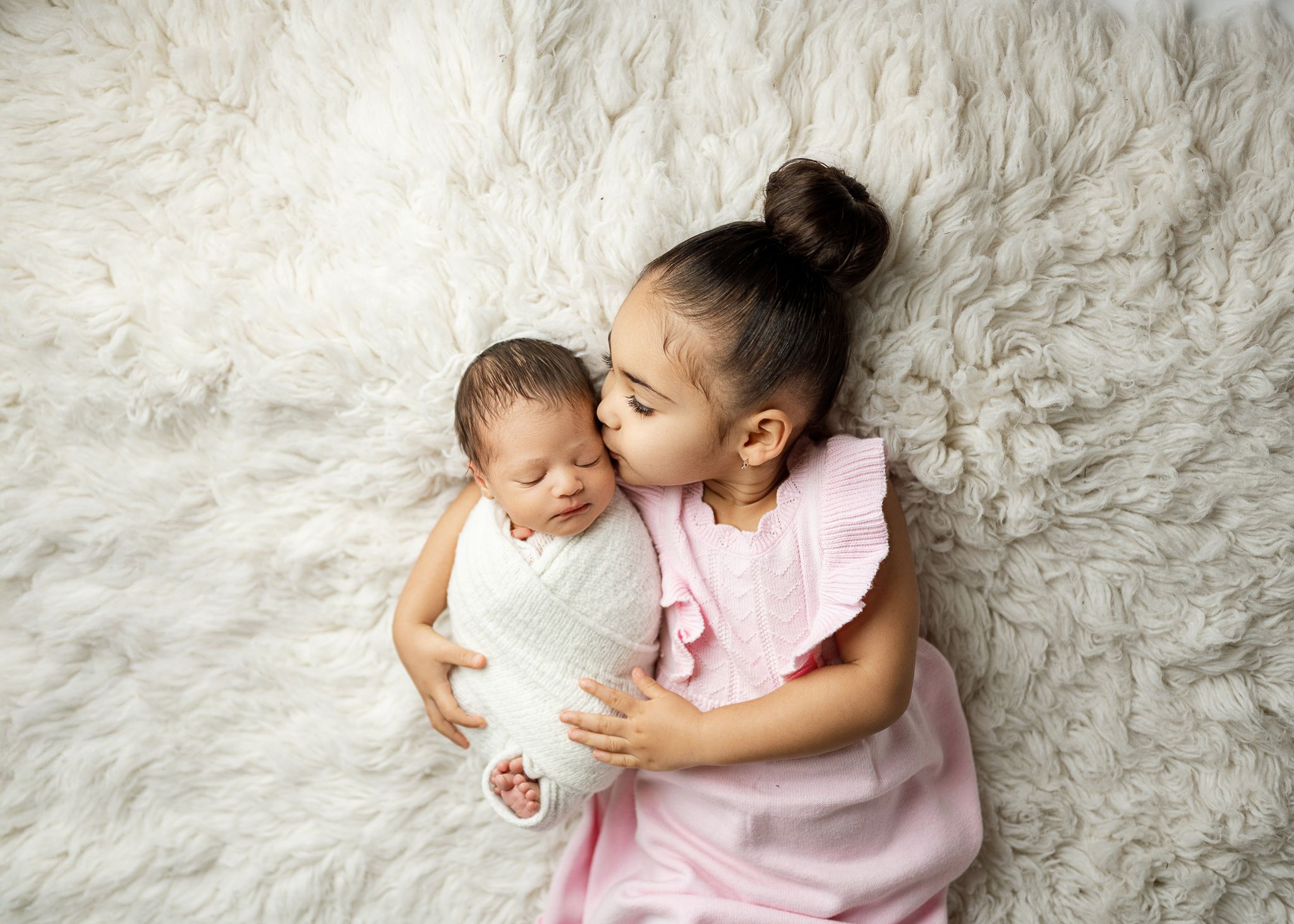 Young child in pink dress tenderly holding and kissing infant sibling while laying on soft white fur rug.