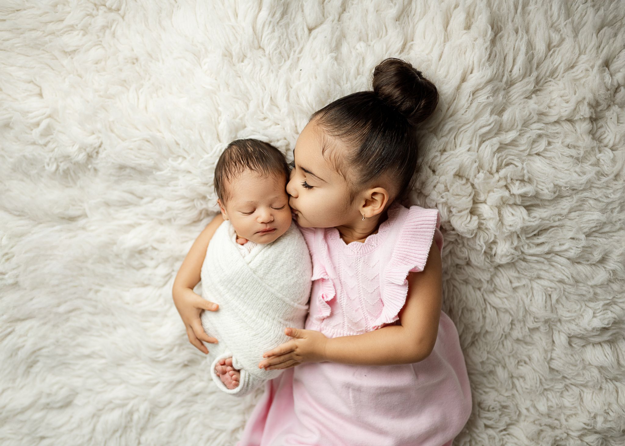 Two young siblings share a tender moment while lying on a soft white fuzzy blanket.