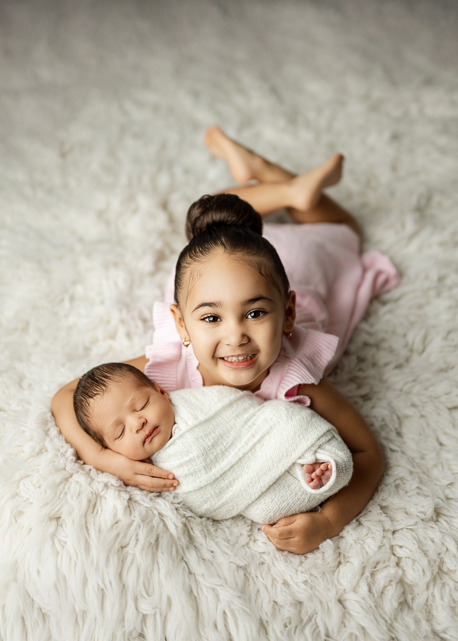 A young child in pink lies on a white fluffy carpet while holding a swaddled newborn baby.