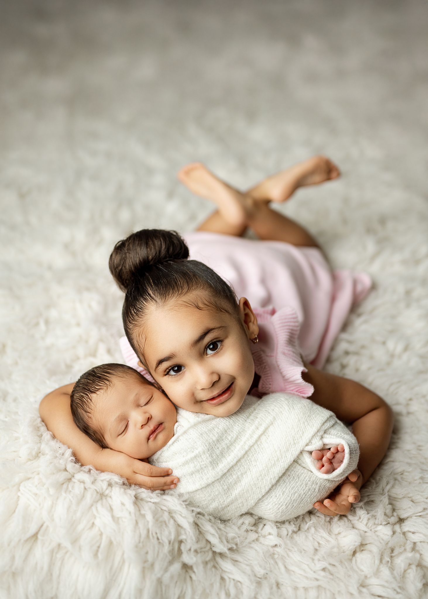 A child and swaddled newborn baby lying together on a white fluffy carpet, sharing a sweet bonding moment.