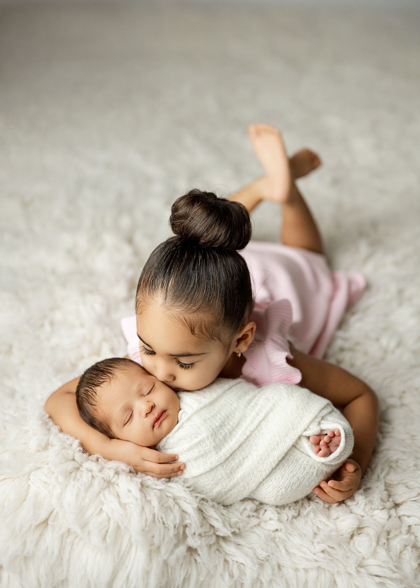 A tender sibling moment shows a child in pink gently embracing a swaddled baby on a fluffy white rug.