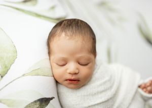 A peaceful sleeping newborn swaddled in soft white fabric against a light floral background.