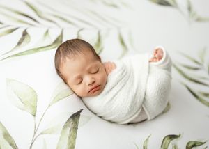 Peaceful sleeping newborn wrapped in soft white swaddle blanket on botanical print background.