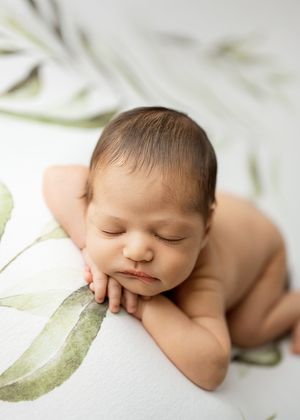 Peaceful sleeping newborn resting with hands under chin on white backdrop with delicate green leaf pattern.
