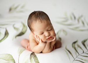 Sleeping newborn lies peacefully on white backdrop with soft green botanical accents in dreamy portrait setting.