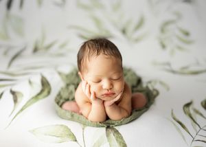 Sleeping newborn rests on elbows with hands under chin on soft green blanket against floral background.