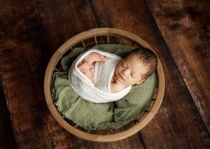 Newborn baby sleeps peacefully in round wooden bowl with soft green fabric and white wrap on dark wooden surface.
