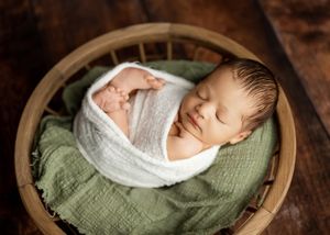 A sleeping newborn baby wrapped snugly in white fabric lies on a textured green blanket in a professional portrait setting.