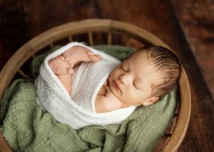 Sleeping newborn wrapped in white swaddle lying peacefully in wooden bowl with green blanket.