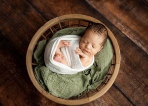 A sleeping newborn wrapped in white fabric lies in a round wooden bowl with soft green textured fabric.