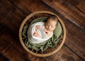 Sleeping newborn baby swaddled in white wrap rests in round wooden bowl with green leaves on rustic wooden floor.