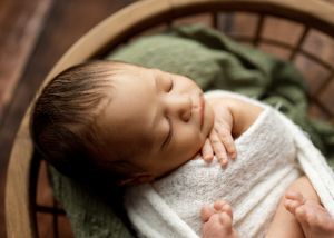 A sleeping baby wrapped in a white blanket rests peacefully in a wooden bassinet with a green cushion.