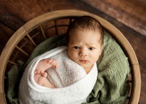 A small baby wrapped in a white swaddle blanket rests in a round wooden basket lined with a soft green cushion.