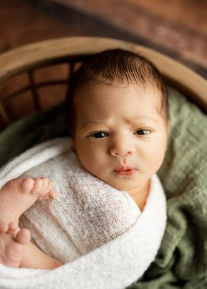 A sleeping newborn swaddled in white fabric lies next to sage green textiles in a peaceful portrait series.