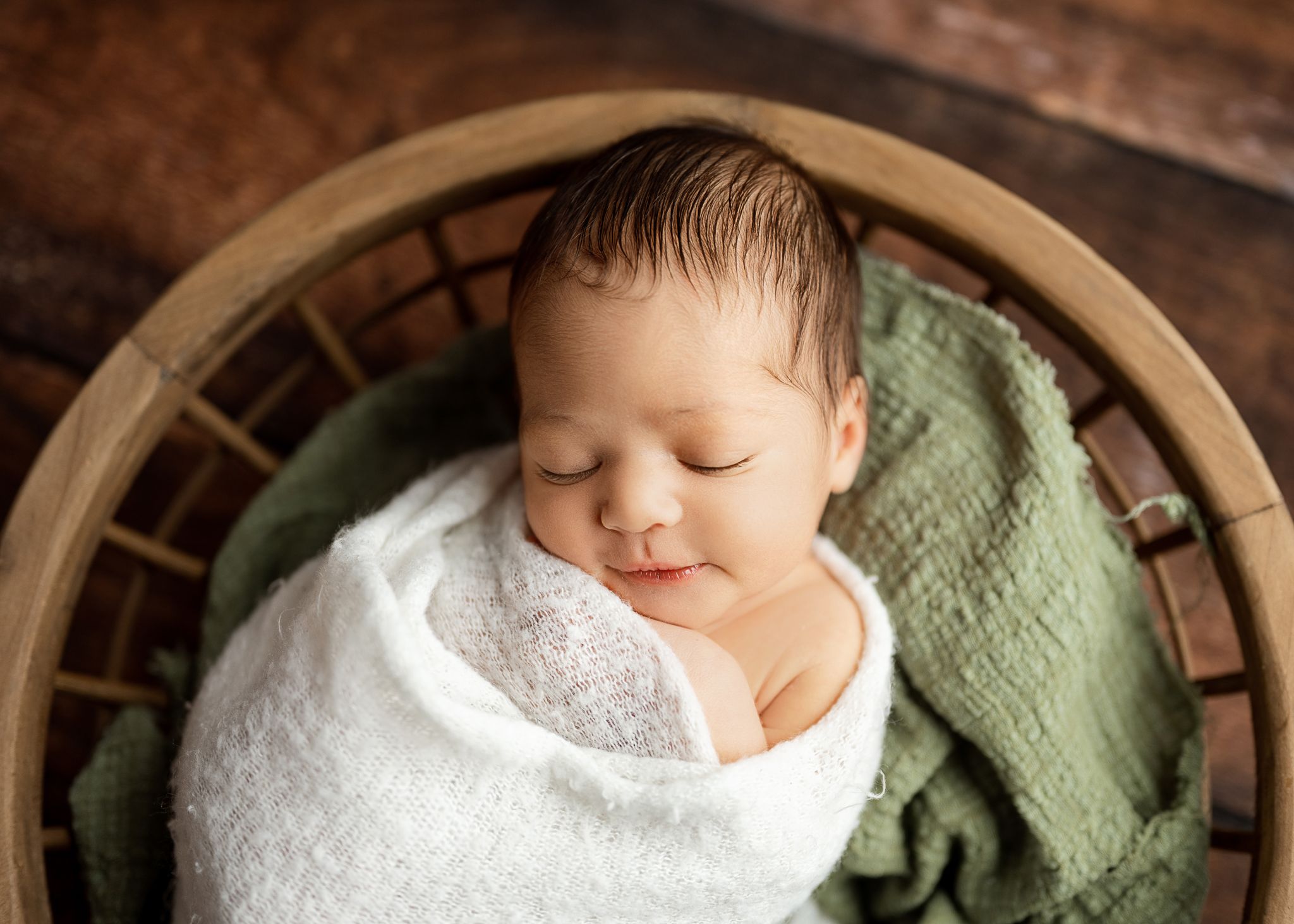 Sleeping newborn baby wrapped in white blanket rests peacefully in wooden bowl with green fabric.