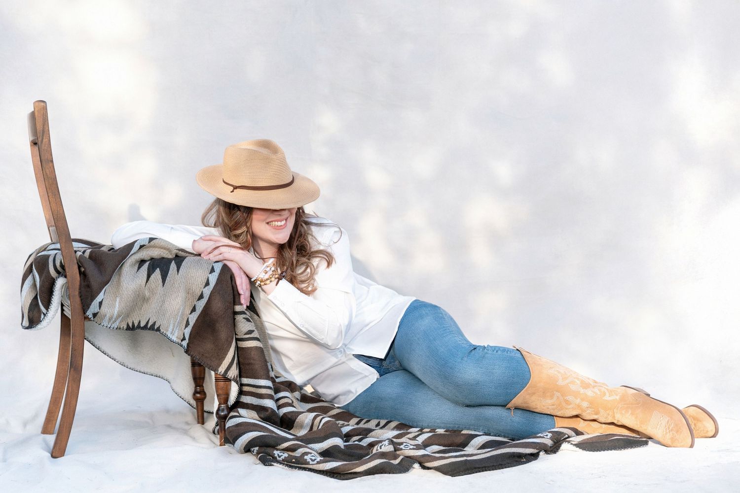 Person in winter attire reclining in snow with skis and wearing a beige hat, white sweater and blue pants.