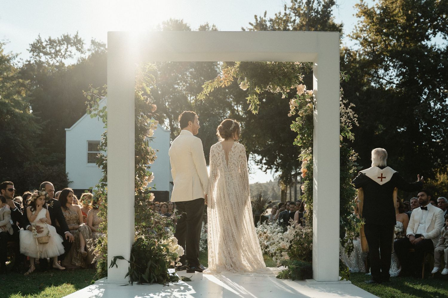 Outdoor wedding ceremony at sunset with white arch and gathered guests beneath sun-dappled trees.