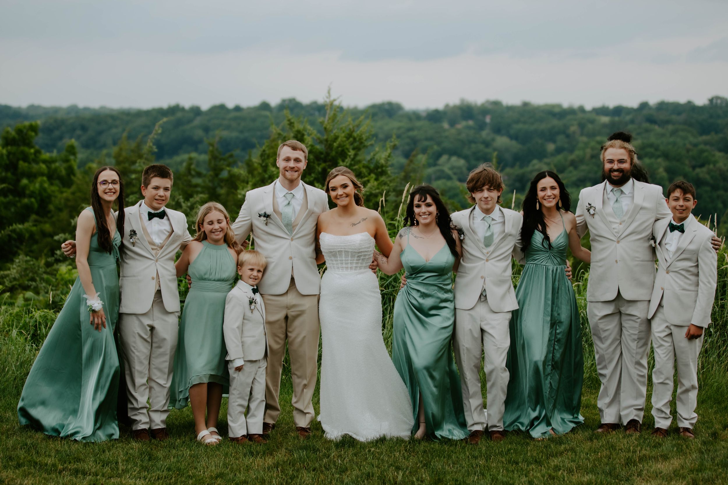 Wedding party group photo with bridesmaids in teal dresses and groomsmen in light gray suits outdoors.