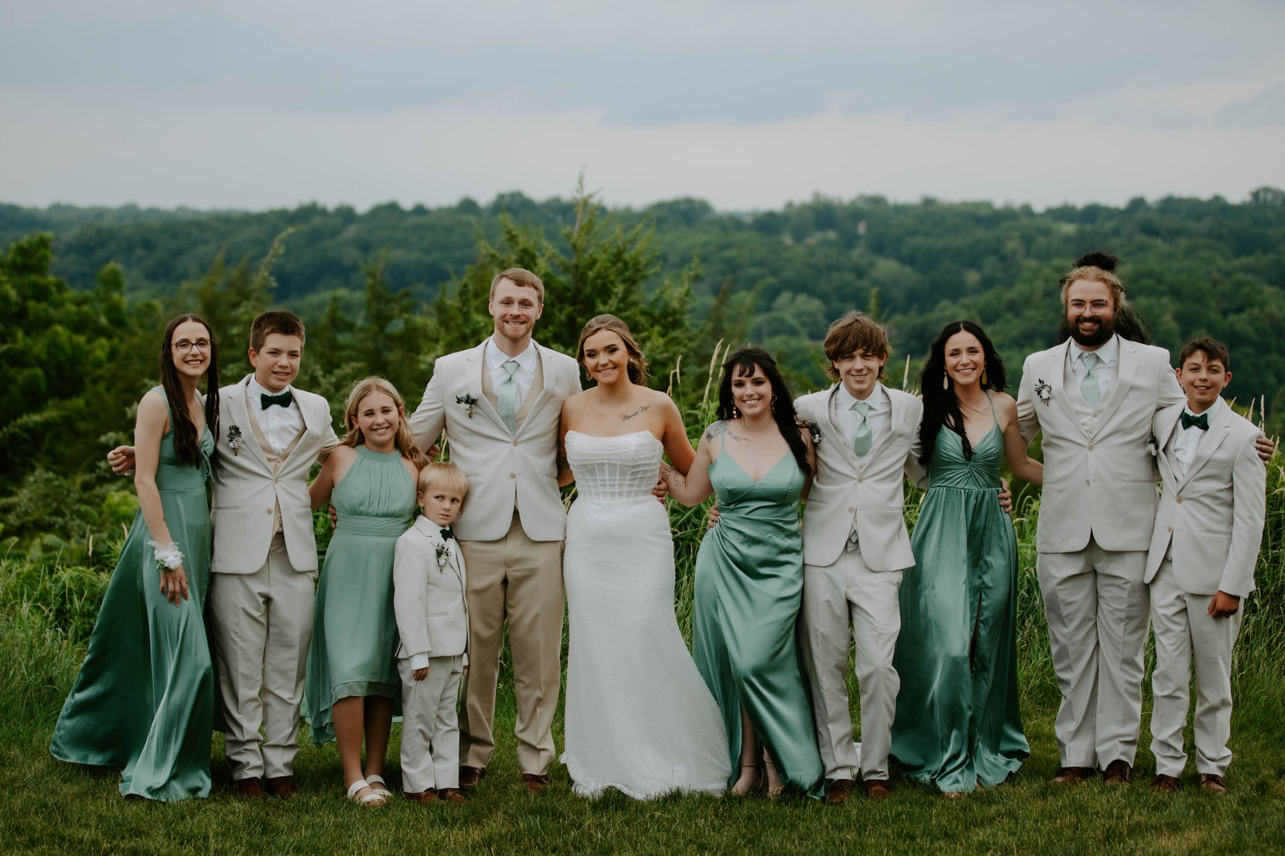 Panoramic wedding party portrait with bridesmaids in teal gowns and groomsmen in light suits against green hills.