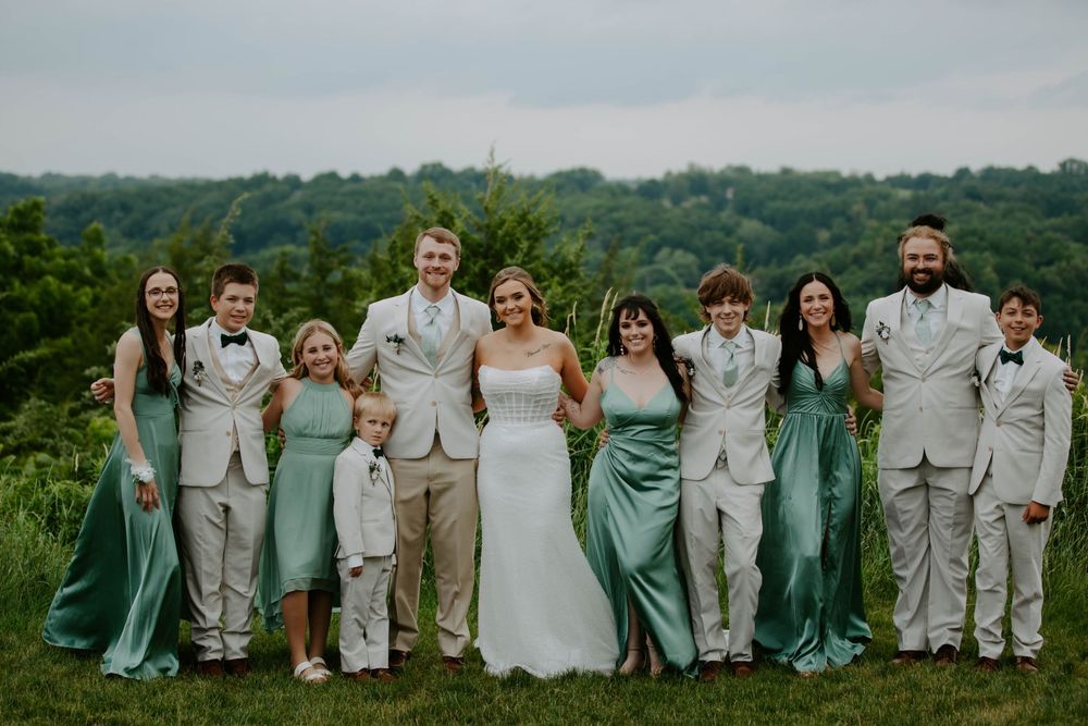 Panoramic wedding party portrait with bridesmaids in teal gowns and groomsmen in light suits against green hills.