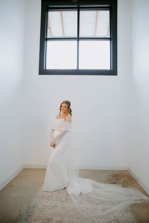 Bride in flowing white dress poses gracefully near window in minimalist white room.