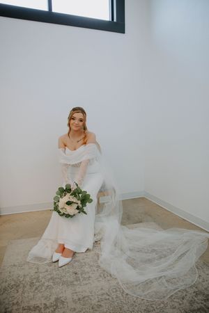 Bridal portrait in a minimalist white room showcasing a flowing wedding gown and bouquet.