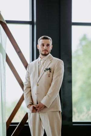 Groomsmen in cream colored suits pose near large windows in a modern venue.
