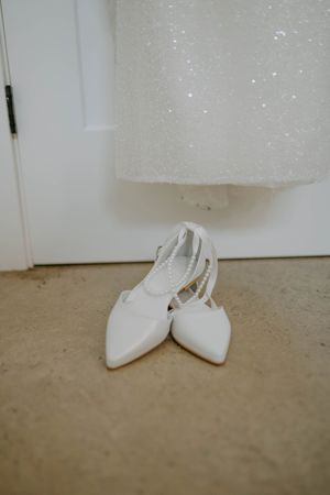Close-up of white bridal shoes on concrete floor with sparkly dress hem visible.