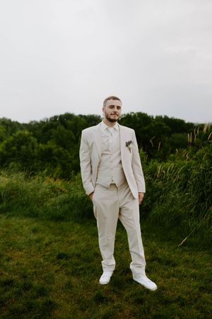 Groom in an all-white suit poses on grassy hillside against cloudy sky.